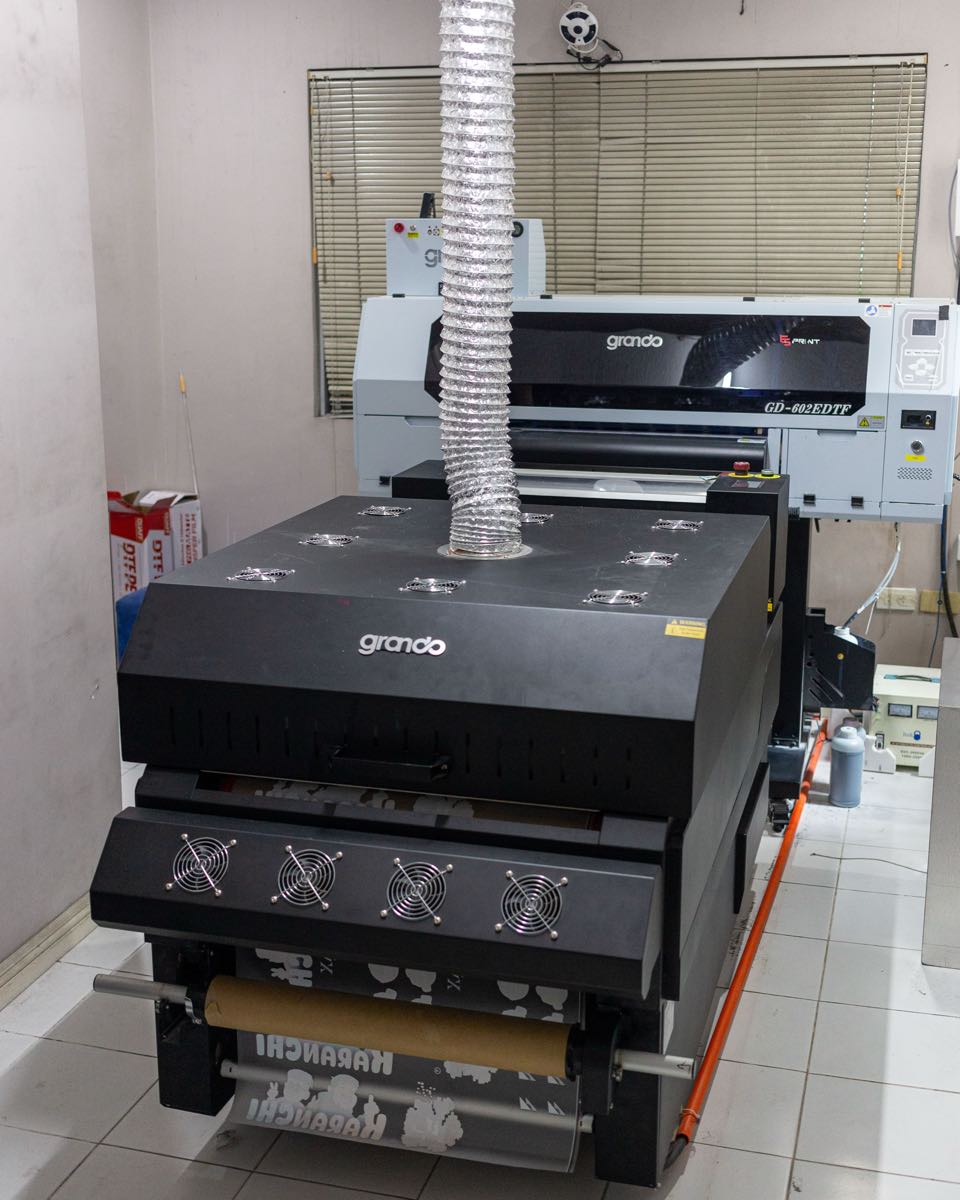 A direct-to-film (DTF) printer and its powder and curing unit in the print room, with a fume extraction duct overhead.