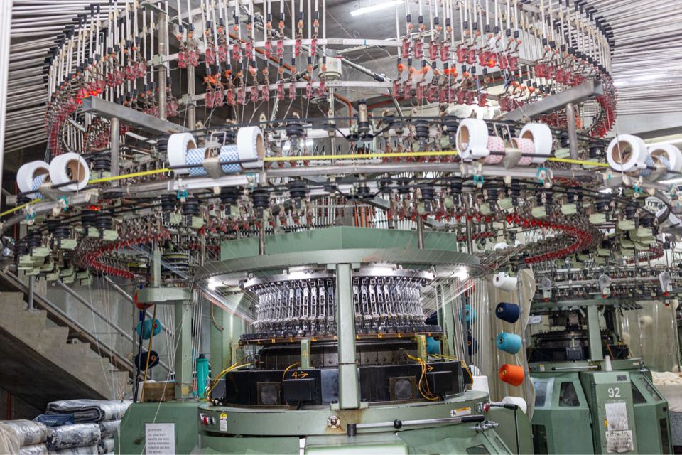 The top of a circular knitting machine, where yarn from a ring of white cones runs down into the feeders around the needle cylinder.
