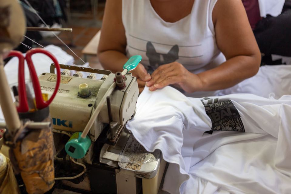 Hands feeding a white garment under the needle of a sewing machine, with red-handled scissors hanging beside it.