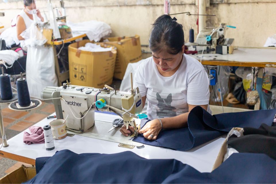 A sewing operator stitches a navy garment panel at an industrial sewing machine, with thread cones and cartons around her.