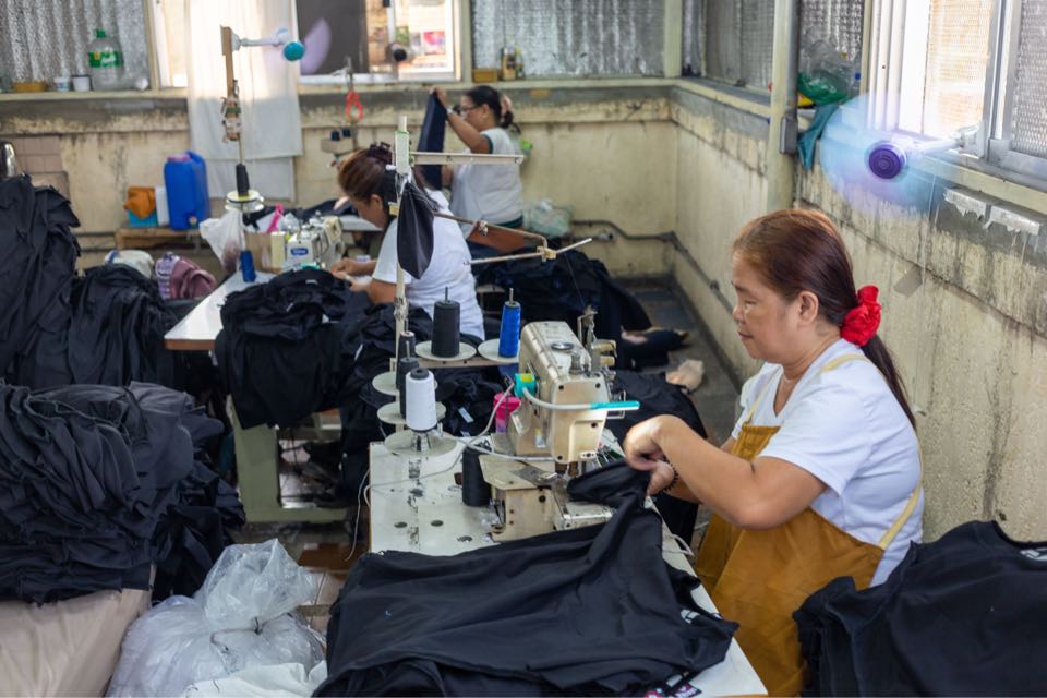 Sewing operators at their machines on the sewing floor, working through piles of black garment pieces.