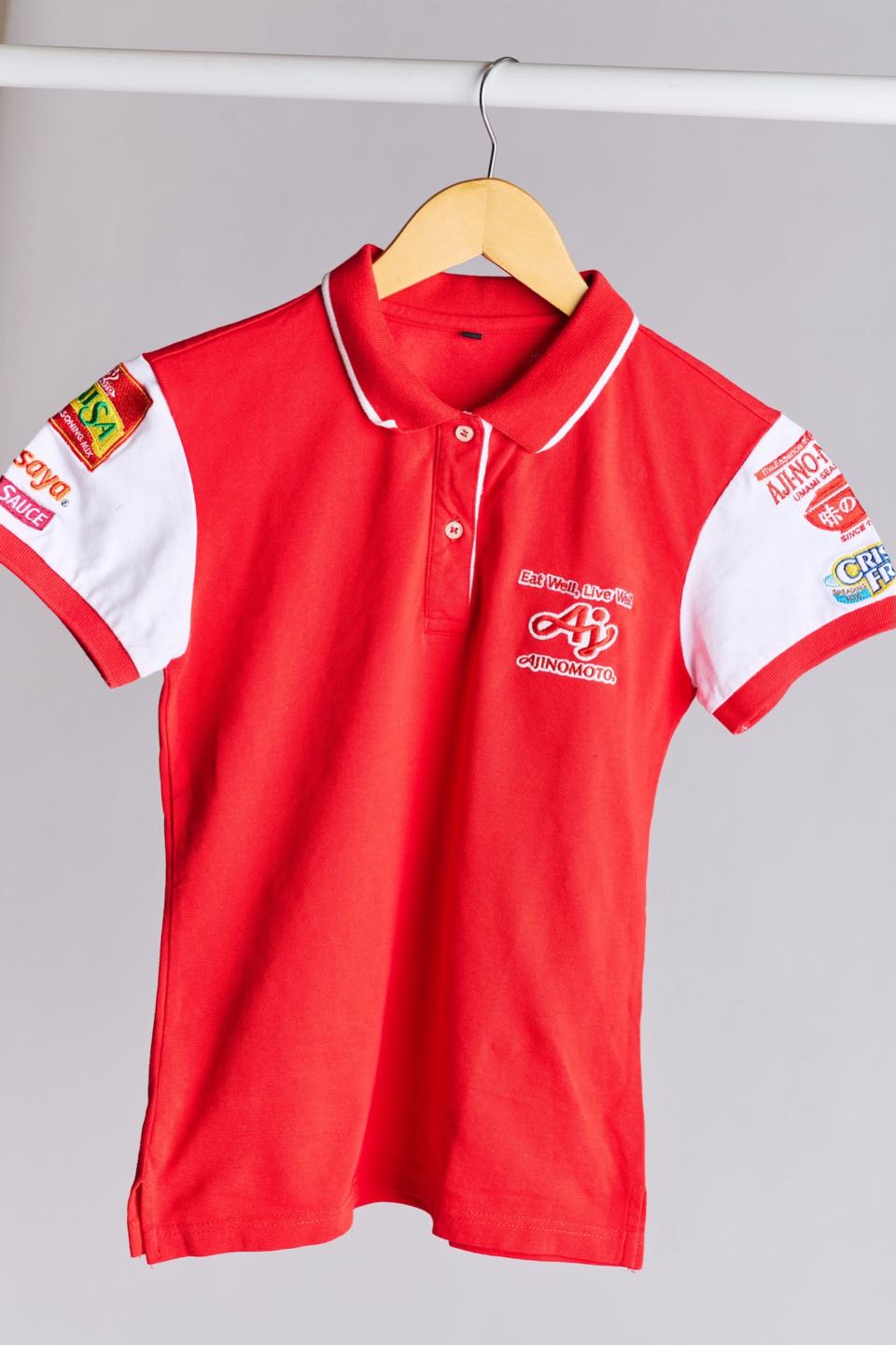 A red Ajinomoto uniform polo on a hanger, with white collar tipping, white sleeve panels and printed brand logos.