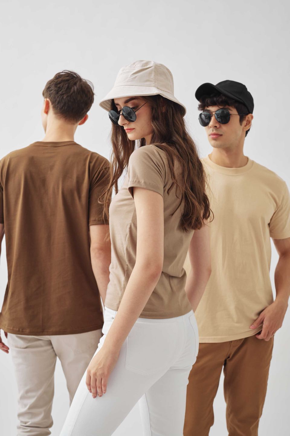 Three models in plain tees in brown, taupe and sand, two of them in sunglasses and caps.