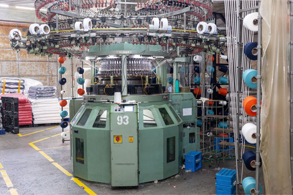A green Fukuhara circular knitting machine, number 93, on the Genknit floor, ringed by cones of colored yarn with wrapped fabric rolls stacked behind it.